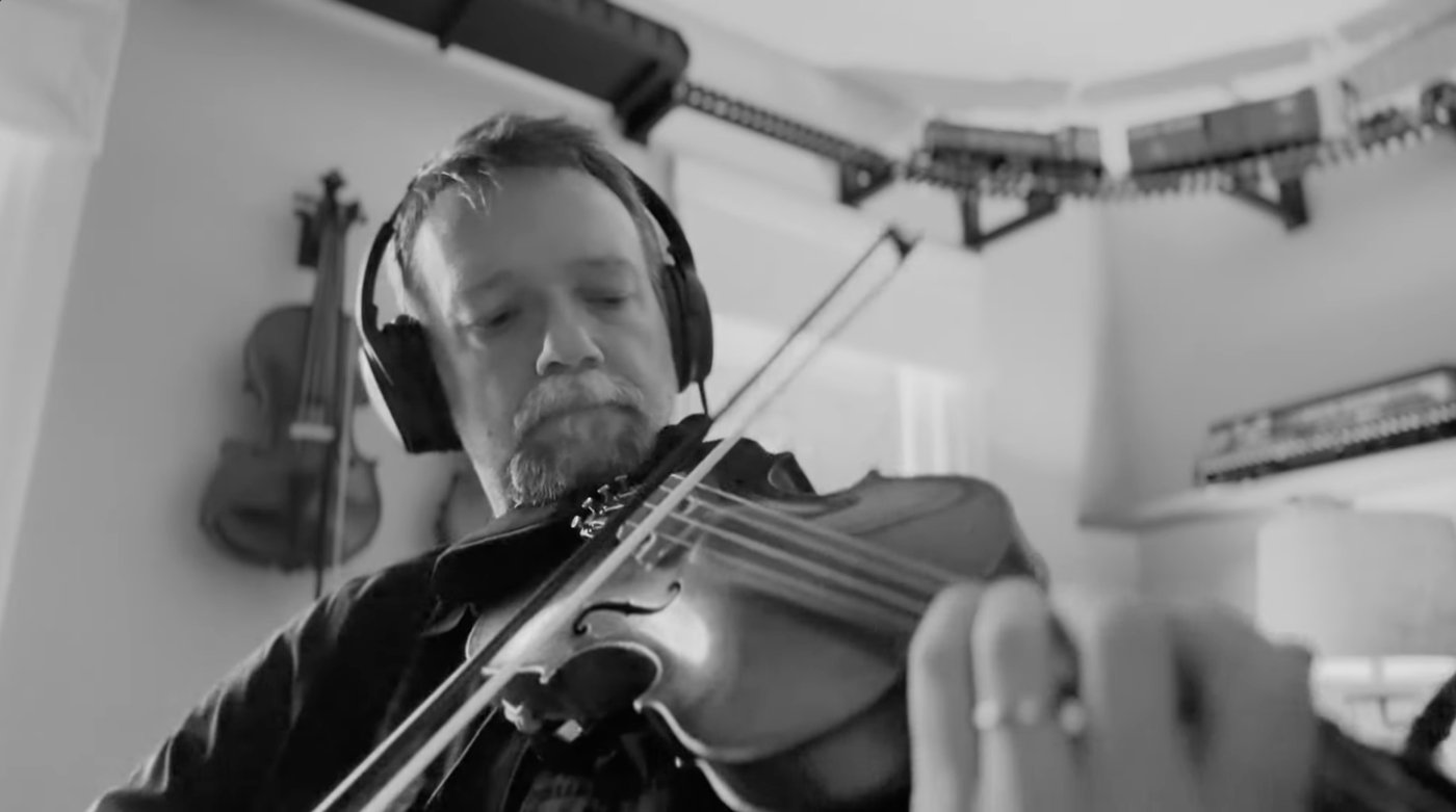 Tod playing the fiddle in his home studio, wearing headphones, with violins hanging on the wall behind him.
